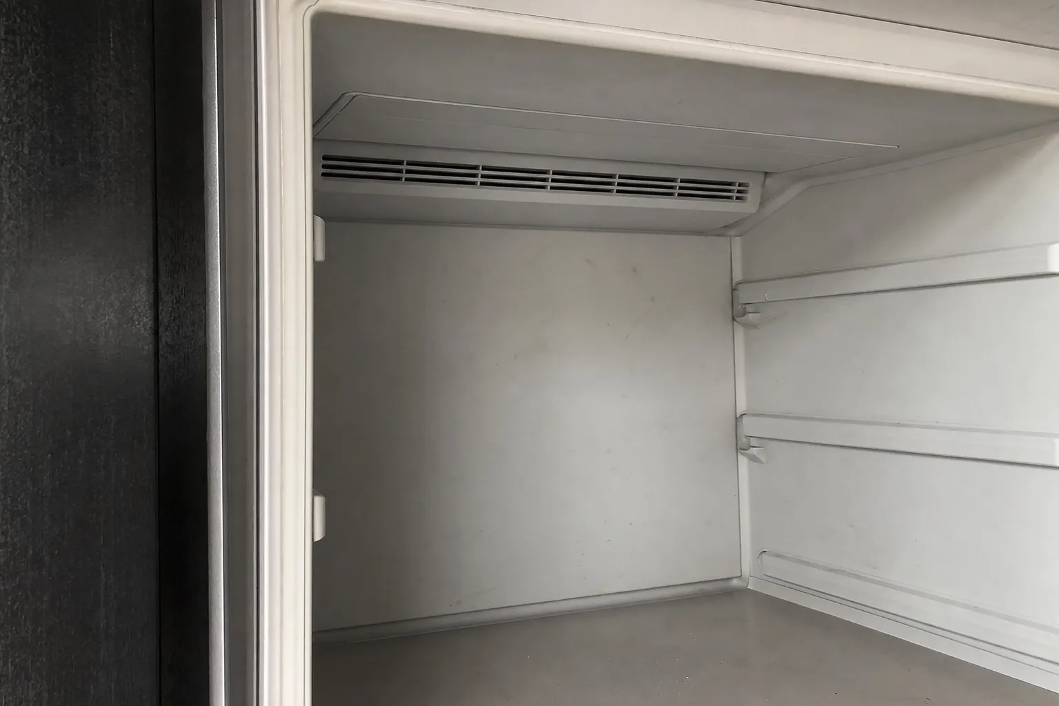 Empty white refrigerator compartment with a slotted air outlet running across the top of the rear wall and bare shelf rails